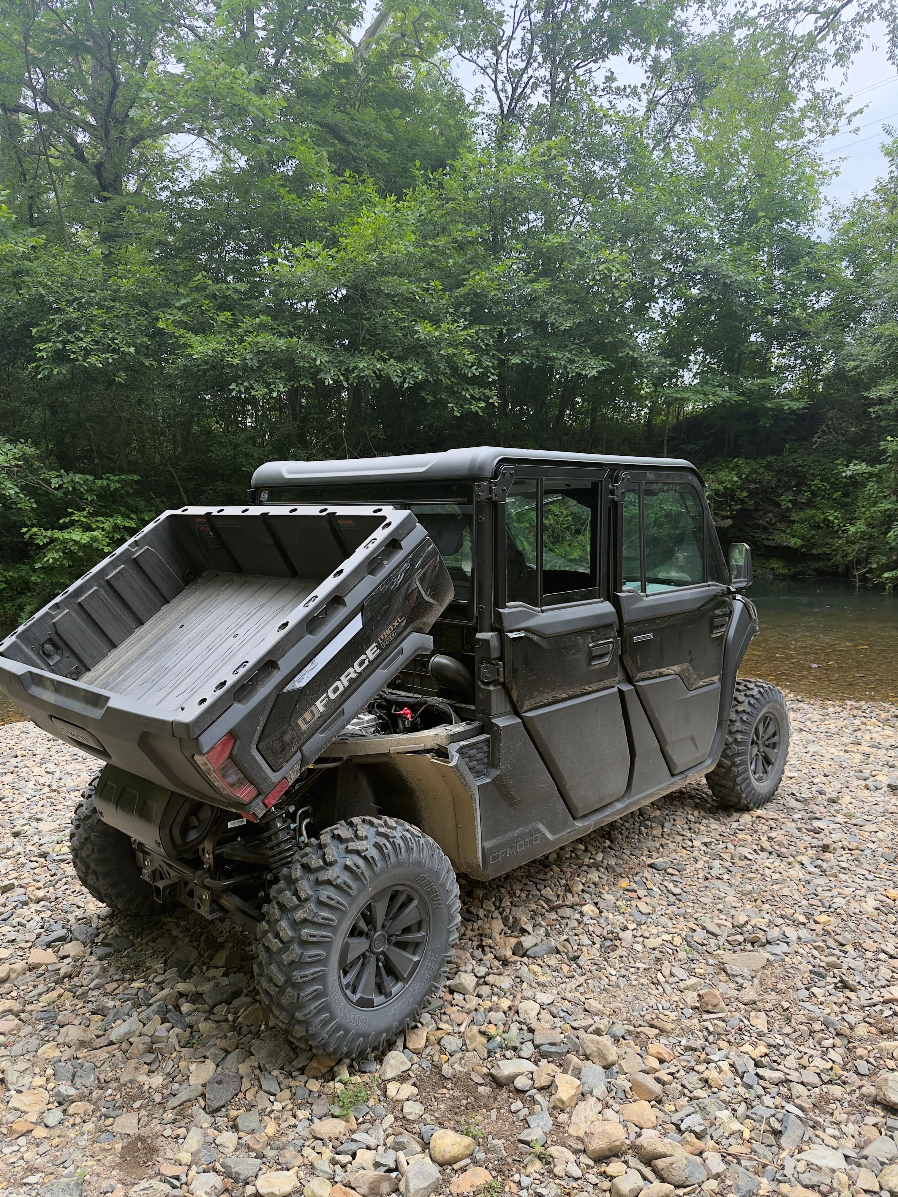 Black four-door UTV with a raised cargo bed on a rocky riverbank near trees.