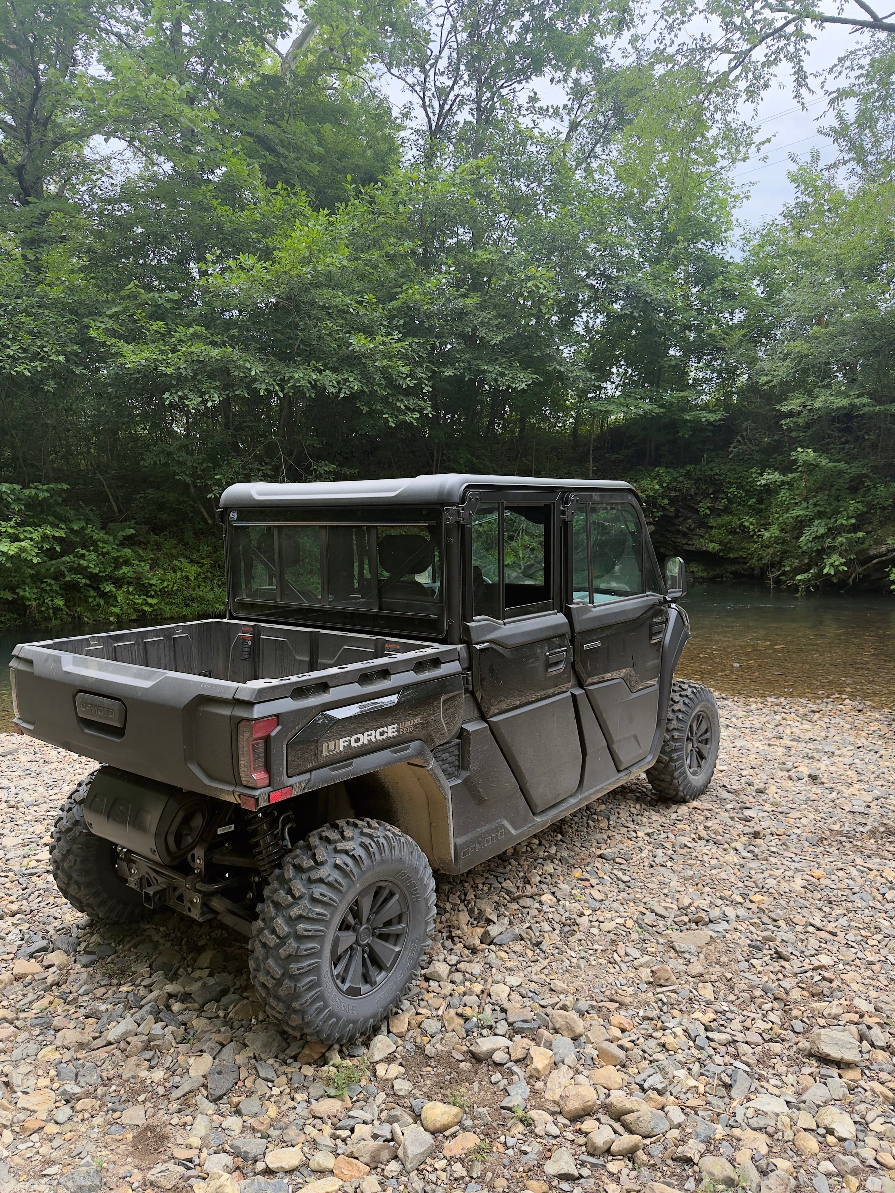 Black four-door CFMOTO UFORCE parked on a rocky riverbank with a dense forest background.