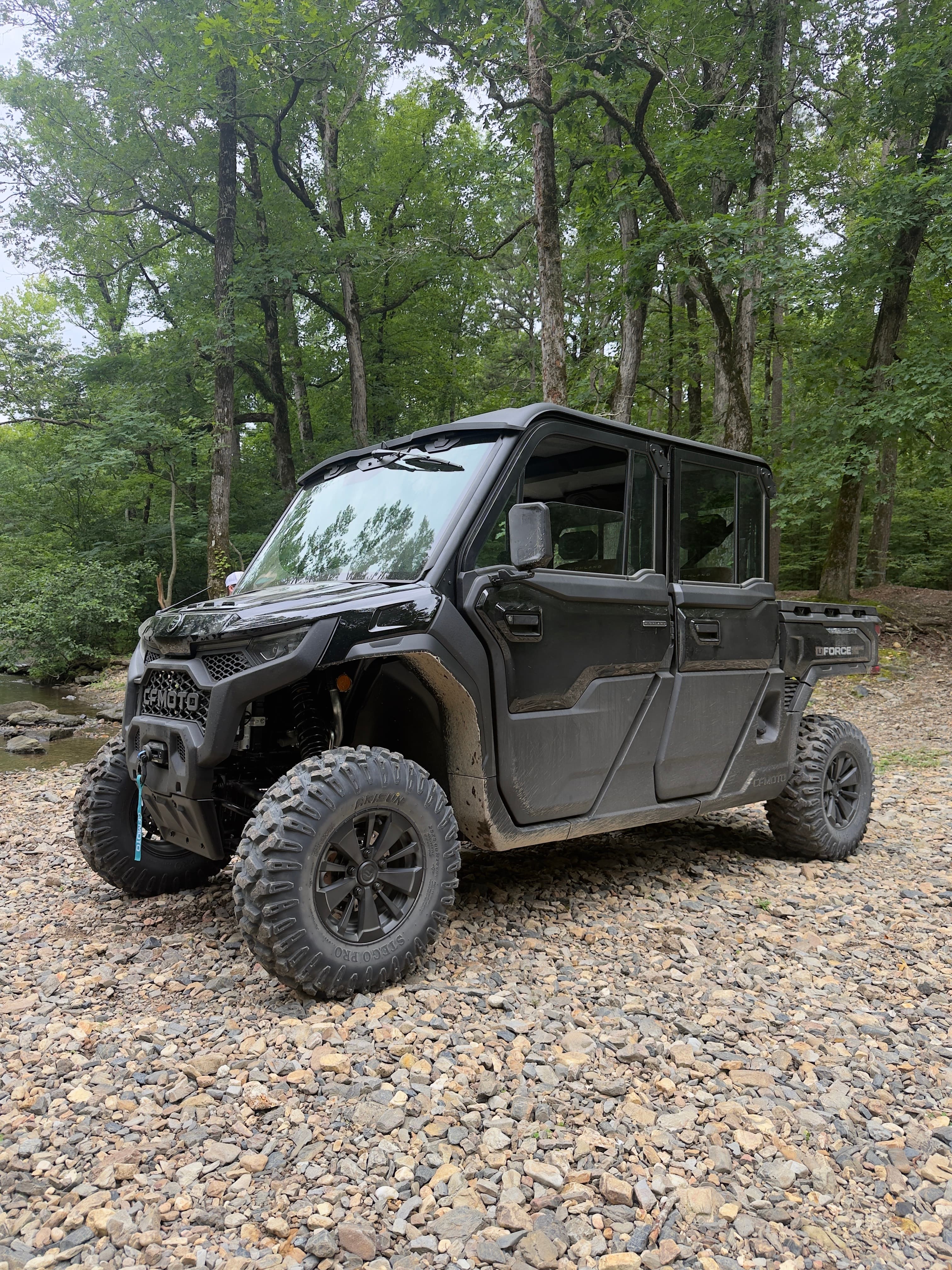 Black CFMOTO UFORCE utility vehicle parked on a rocky riverbank with a dense forest background.
